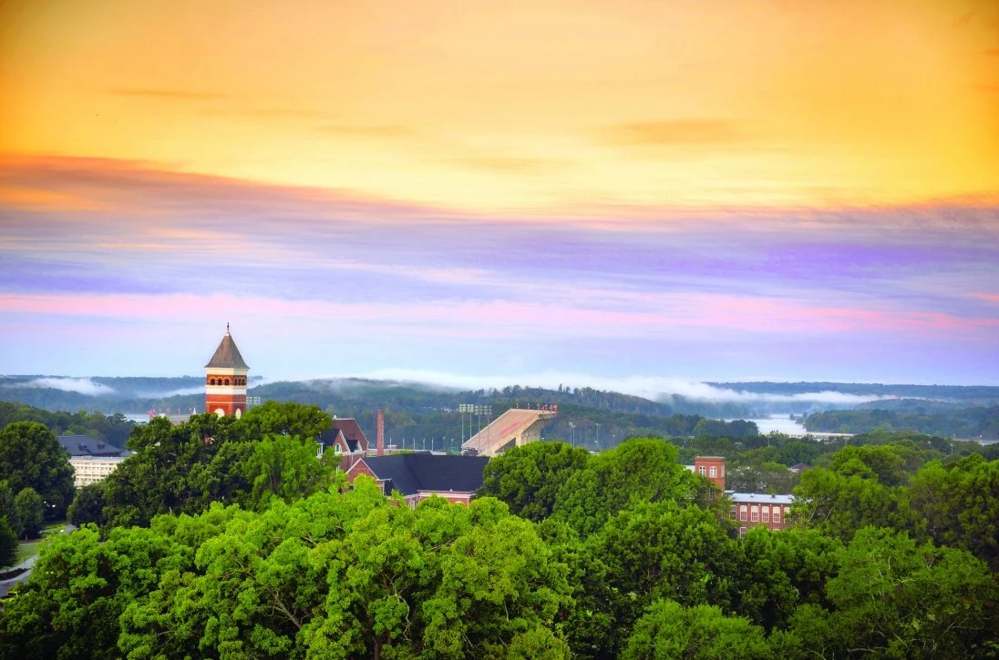 Aerial view of a green, tree-covered campus with a brick tower and distant stadium under a colorful sunset sky